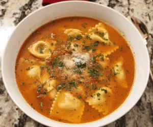 A bowl of hearty ravioli soup featuring stuffed ravioli, fresh vegetables, and herbs in a flavorful broth, served garnished with basil and grated cheese.